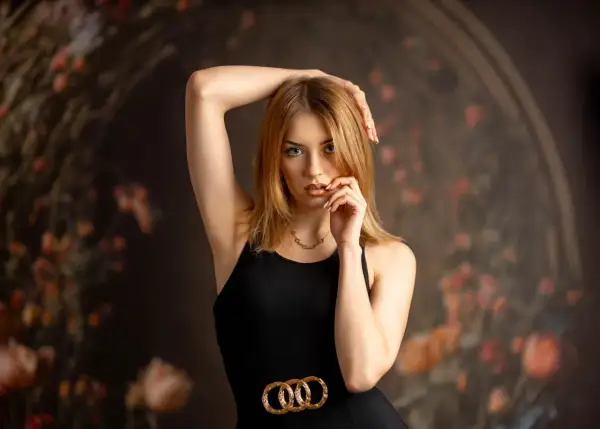 Zmysłowe zdjęcie modelki w czarnej sukience z złotym paskiem, z ręką przy twarzy.